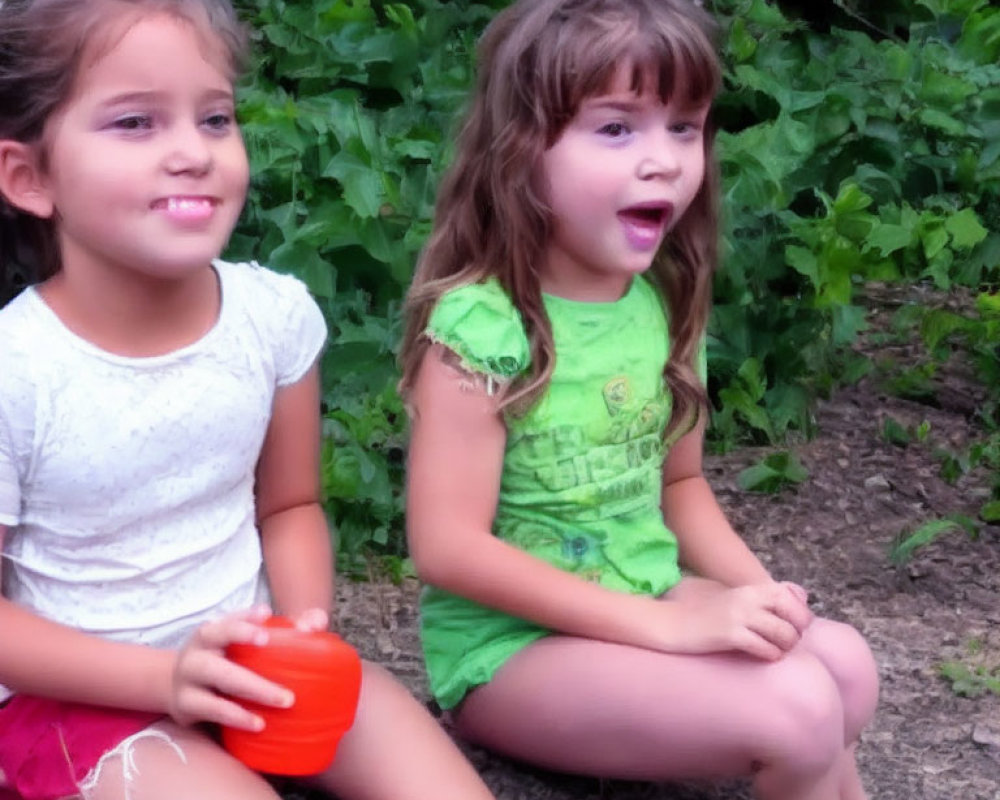 Two young girls sitting outside, one in green shirt with excited expression, the other smiling slightly.