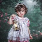 Woman in whimsical attire with bow holding vintage clock in dreamy forest with purple flowers.