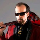 Elderly man in sunglasses plays double-neck guitar in red and black outfit under cloudy sky