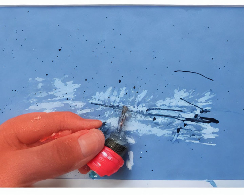 Hand squeezing black paint onto brush on blue paper with splatters