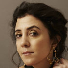 Dark-haired woman with subtle makeup in neutral expression on tan backdrop