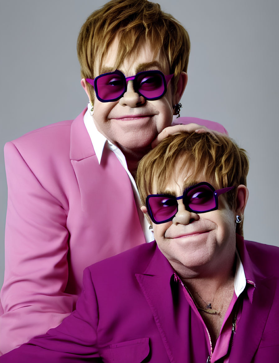 Two people in pink outfits and purple glasses pose against grey backdrop