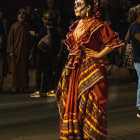 Person in Vibrant Orange Dress with Skull Mask and Floral Headpiece Surrounded by Shadowy Figures with