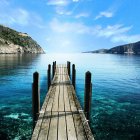 Tranquil lakeside view with wooden dock, houses on stilts, mountains, and sailboat