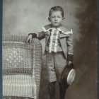 Vintage-style portrait of young boy in formal attire by tree with bellhop's bell, against muted backdrop