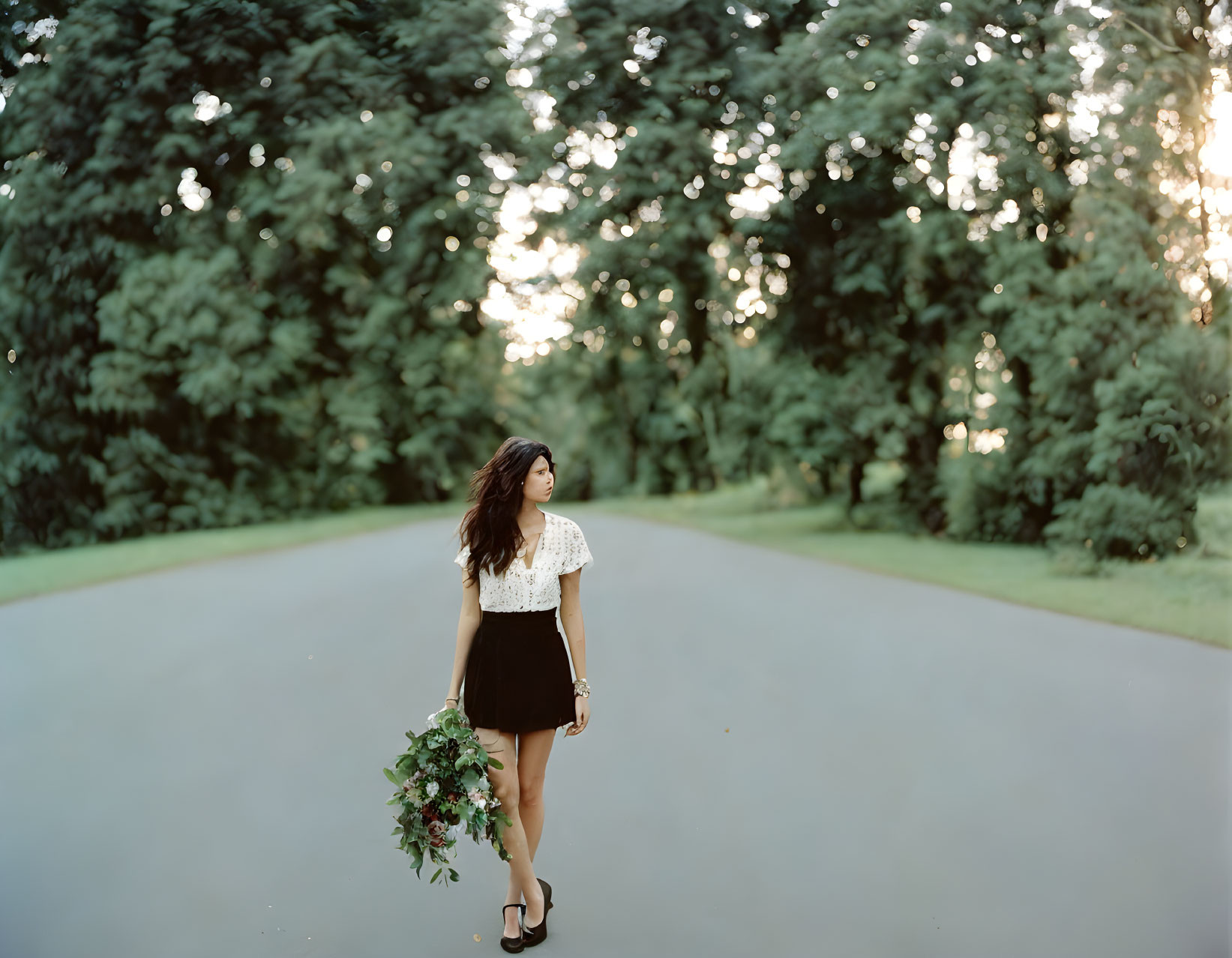 Woman in white blouse and black skirt holding flowers on a road with trees and dappled sunlight.