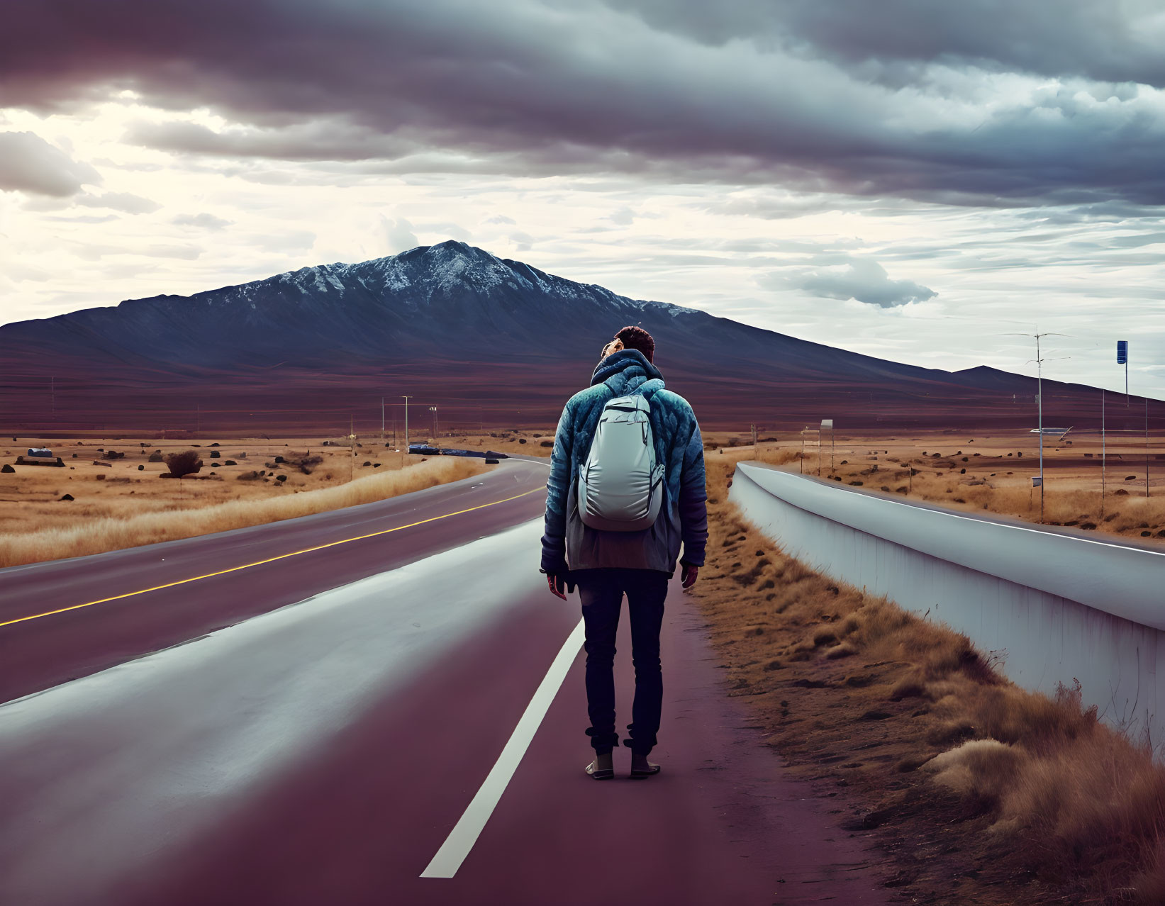 Backpacker walking on empty road towards mountain under cloudy sky