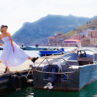 Woman in flowing dress on dock with boats, mountains, and clear skies