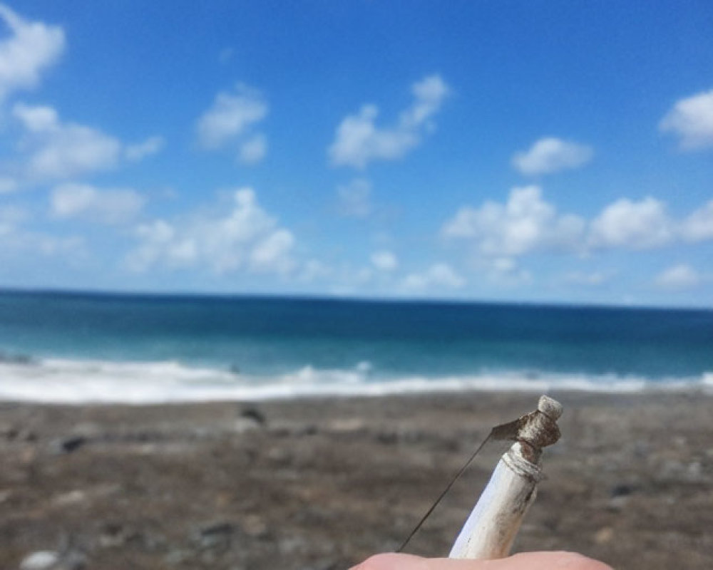 Hand holding cigarette with ash buildup against blue sky, clouds, calm sea