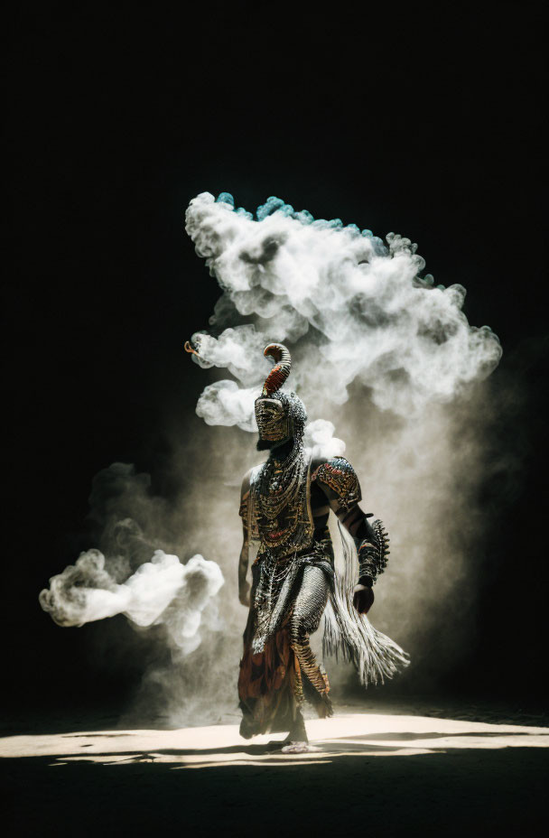 Person in ornate costume with plumed headdress amidst swirling smoke on dark background spotlighted from above