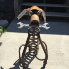 Metal dog sculpture with mechanical parts and white skeletal forelegs outdoors.