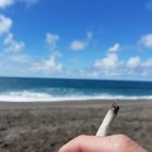 Hand holding cigarette with ash buildup against blue sky, clouds, calm sea