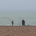 Anglers by shore with boats, rough sea waves, birds, cloudy sky