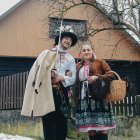 Traditional Andean clothing worn by two people in front of a house with a flowering tree and intricate patterns