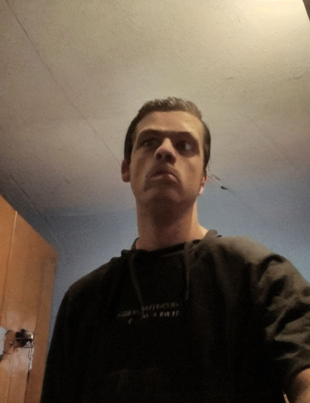 Serious person in black hoodie indoors with ceiling and door.