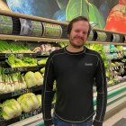 Man with bun and sunglasses in front of grocery store vegetable section with cosmic orbs.