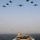 Military aircraft flying over naval formation with aircraft carriers and escort ships in clear sky