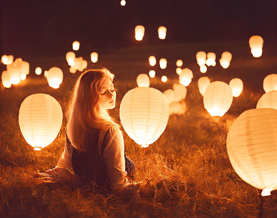 Woman sitting on grass surrounded by glowing paper lanterns at night