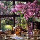 Vintage interior with oil lamp, copper tea set, books, pink blossoms, and garden view.