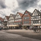 Historic cobblestone village plaza with old buildings, pedestrians, and flower petals-covered river