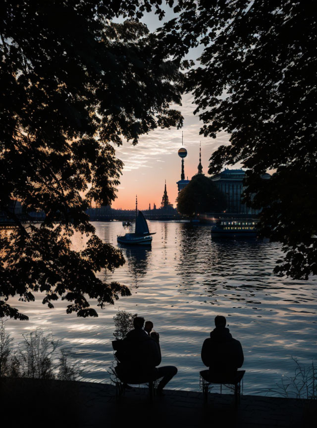 Silhouetted Figures by River at Sunset with Boats and Tower