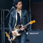 Dark-Haired Person in Metallic Jacket Holds Electric Guitar on Cosmic Background