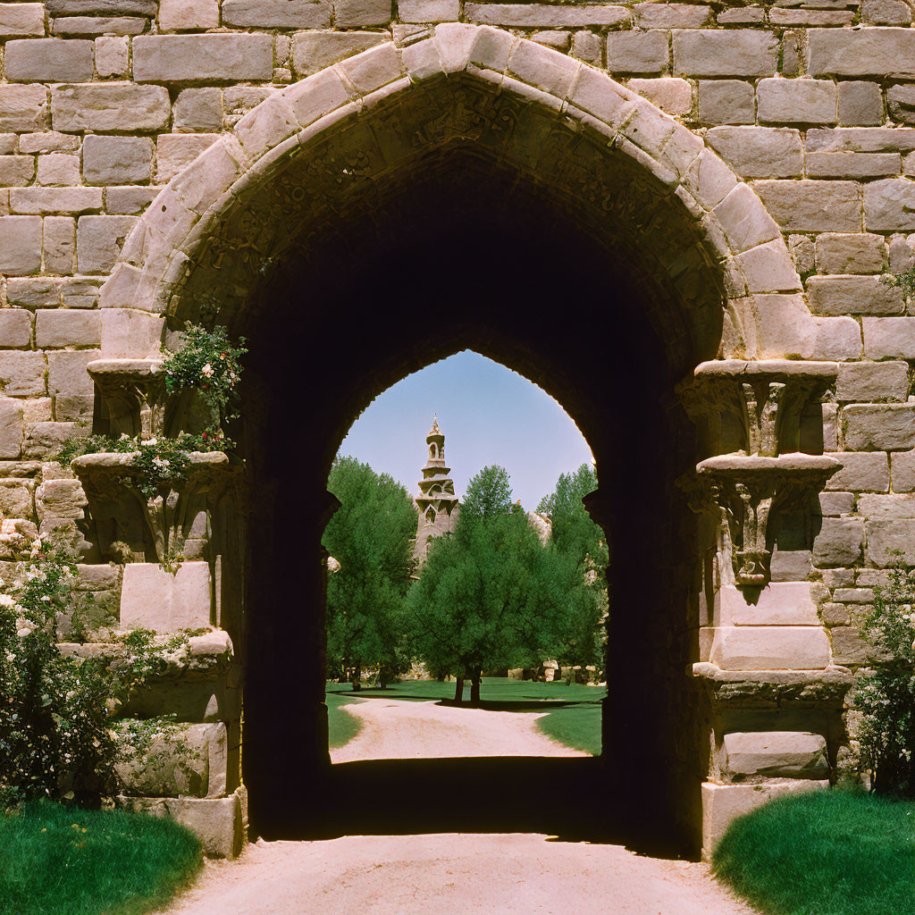 Stone archway frames tree-lined path to distant tower under clear blue sky