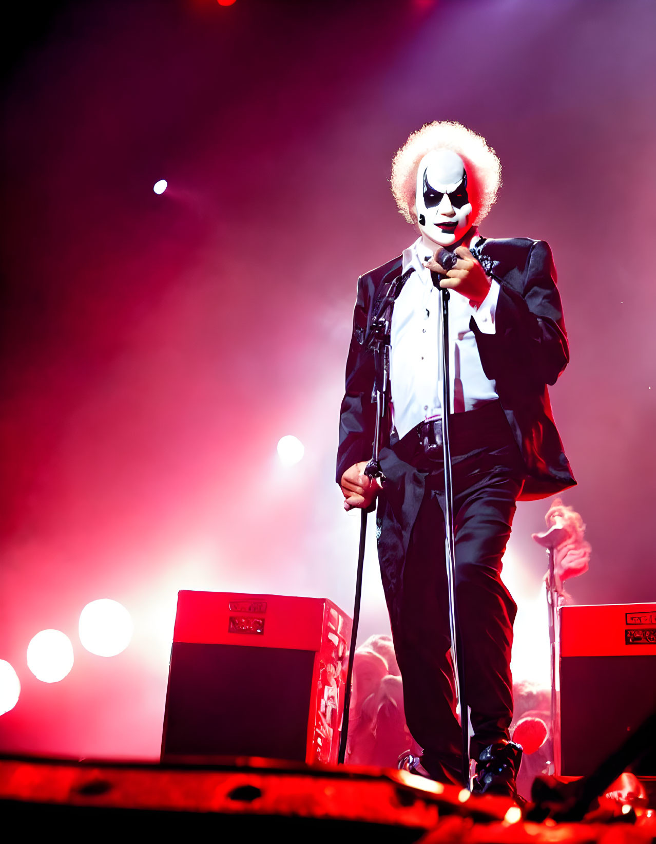 Masked performer in black suit under red lighting on stage with microphone