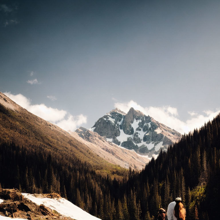 Snow-capped mountain peak overlooking forest with two people gazing up