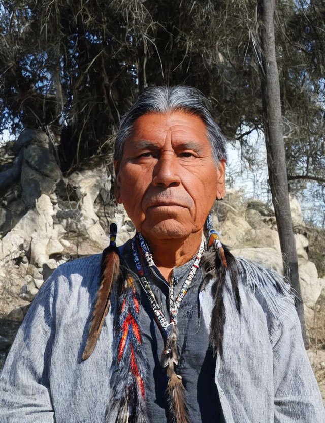 Portrait of stern man with long gray hair in traditional attire and feathered accessories against nature backdrop