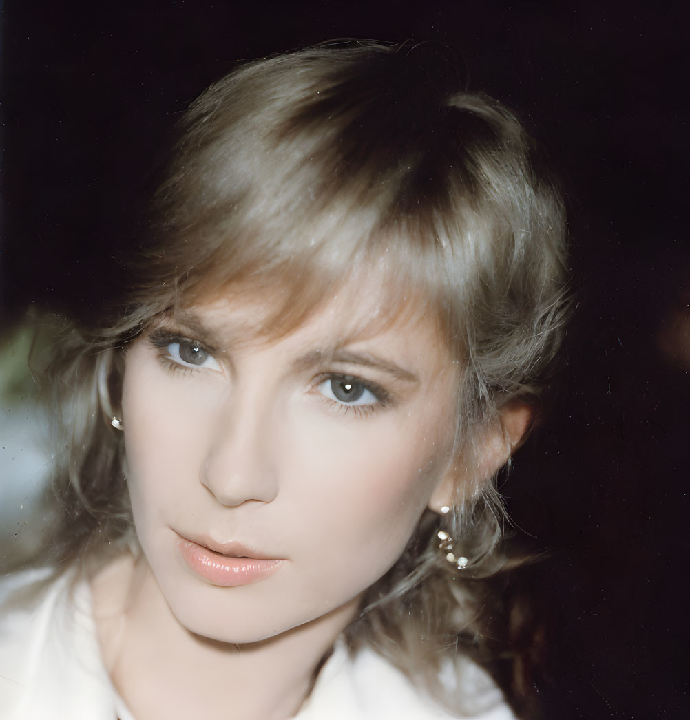 Blonde woman with blue eyes in white top and earrings gazes at camera