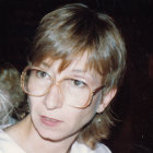 Blonde woman with blue eyes in white top and earrings gazes at camera