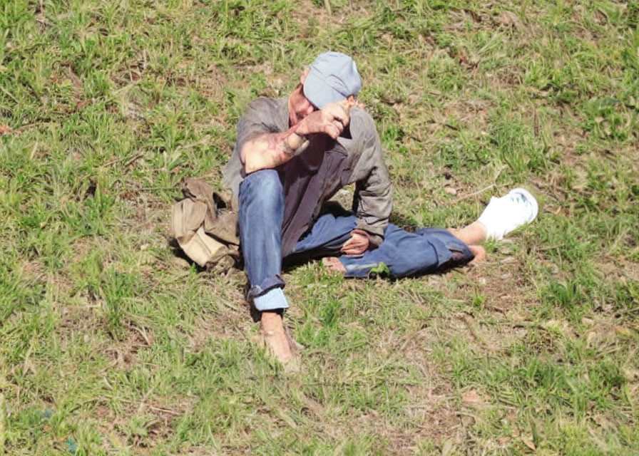 Person sitting on grass wearing cap, covering face with hand, one shoe off.