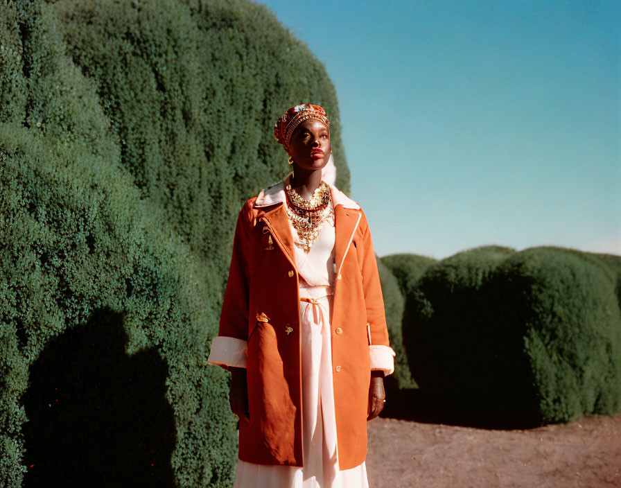 Stylish woman in orange coat and white dress with bold jewelry in garden setting