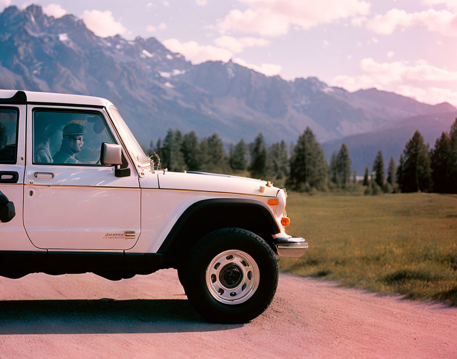 White SUV with driver parked on dirt road amidst mountains and tall grass under pink sky