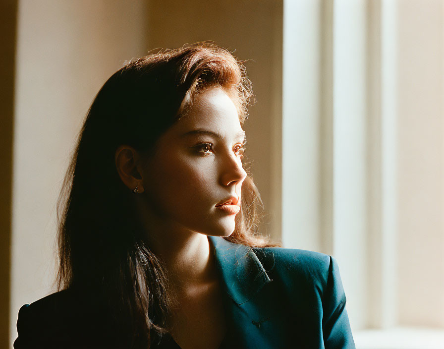 Contemplative woman by window with teal blazer and sunlight shadows