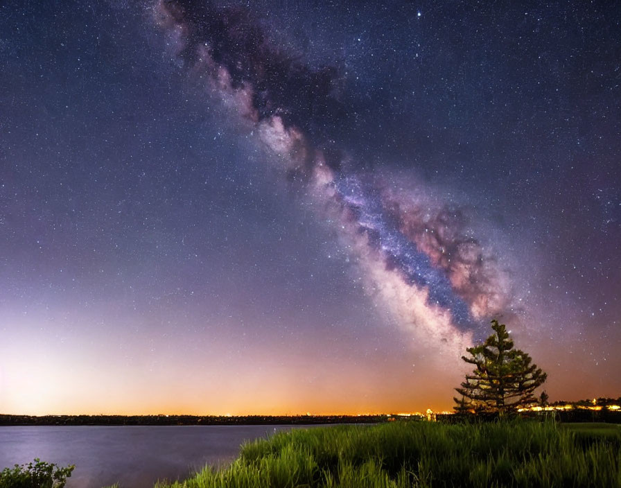 Panoramic night sky with Milky Way over tranquil lake, tree, and grassy shore