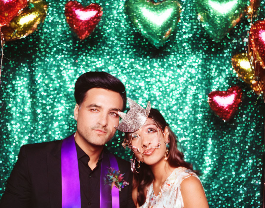 Couple in festive party attire with heart-shaped balloons and sparkling backdrop