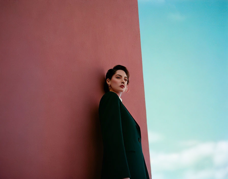 Pensive woman in dark coat against pastel pink wall under soft blue sky