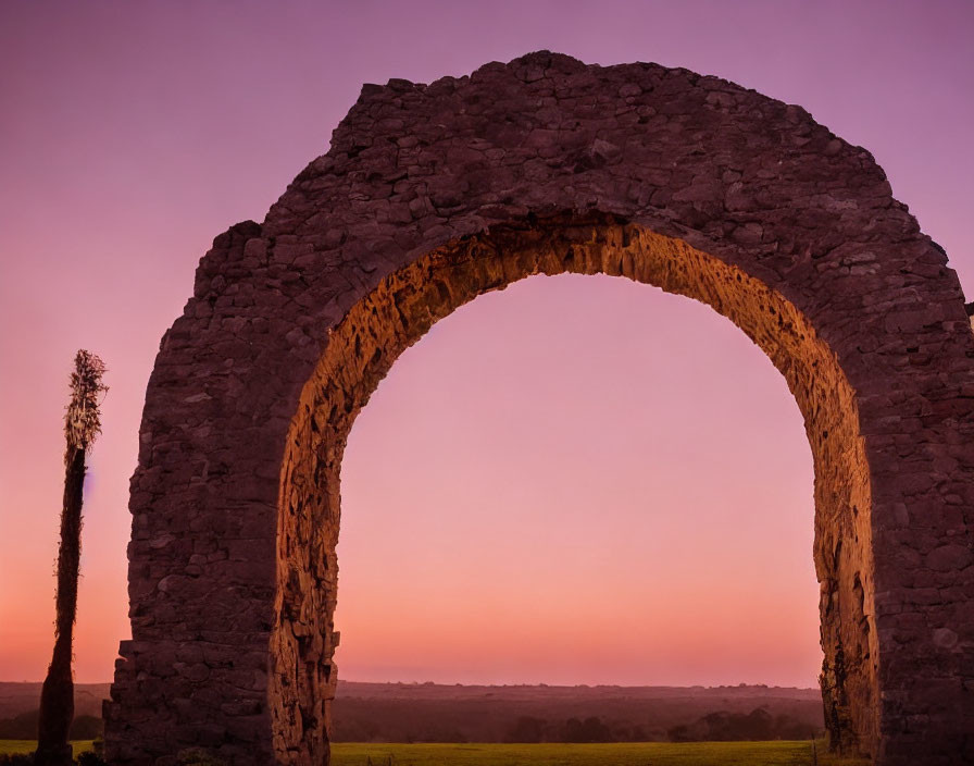 Stone arch against pink and purple sunset sky with lone tree.