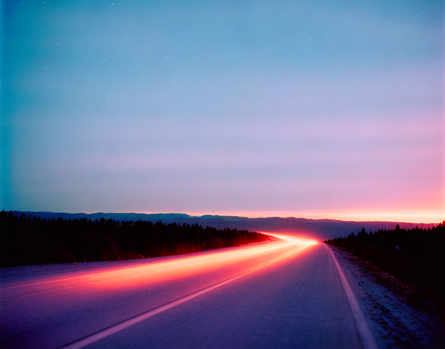 Snowy road at twilight with car tail lights streaking under starry sky