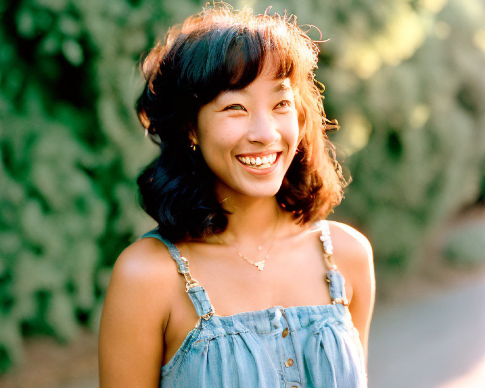 Smiling woman in denim dress outdoors with greenery.