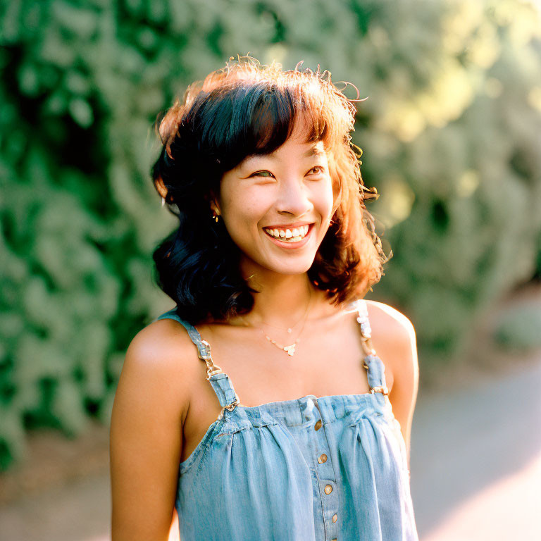 Smiling woman in denim dress outdoors with greenery.