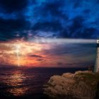 Lighthouse on rocky coastline with red-roofed building at sunset