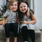 Two girls in fancy dresses smiling on sunlit steps with flowers