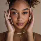 Woman with ornate golden headpiece and metallic tattoos poses with hands framing face