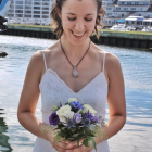 Bride in strapless gown with tiara, holding white and purple bouquet by water and buildings.