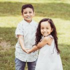 Children in vintage attire holding hands by sunlit pond with flower-adorned hair