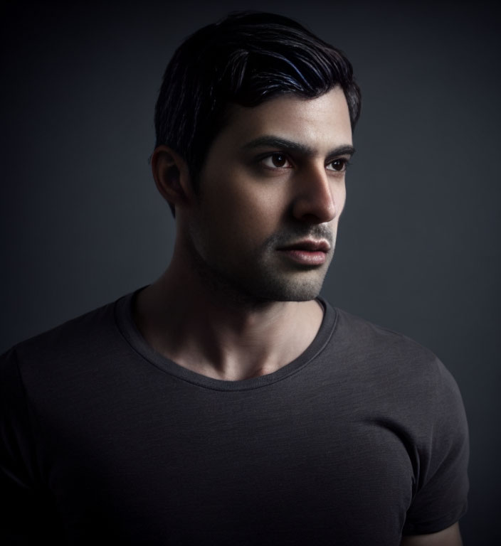 Dark-haired man in black t-shirt with serious expression, looking sideways on dark backdrop.