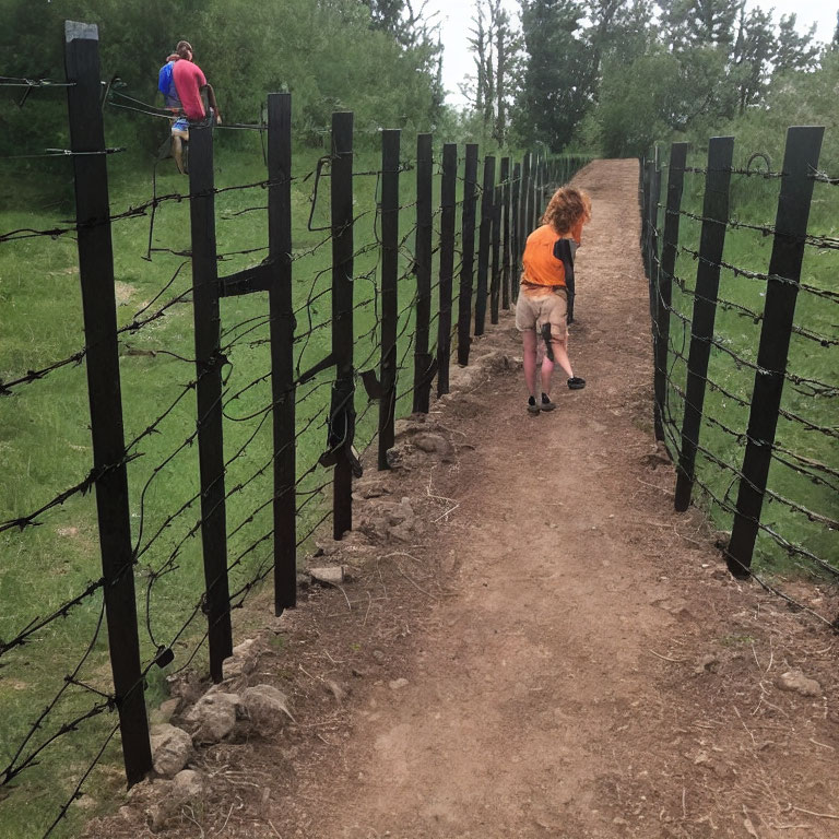 Child in orange shirt walking near barbed wire fence with adult and trees.